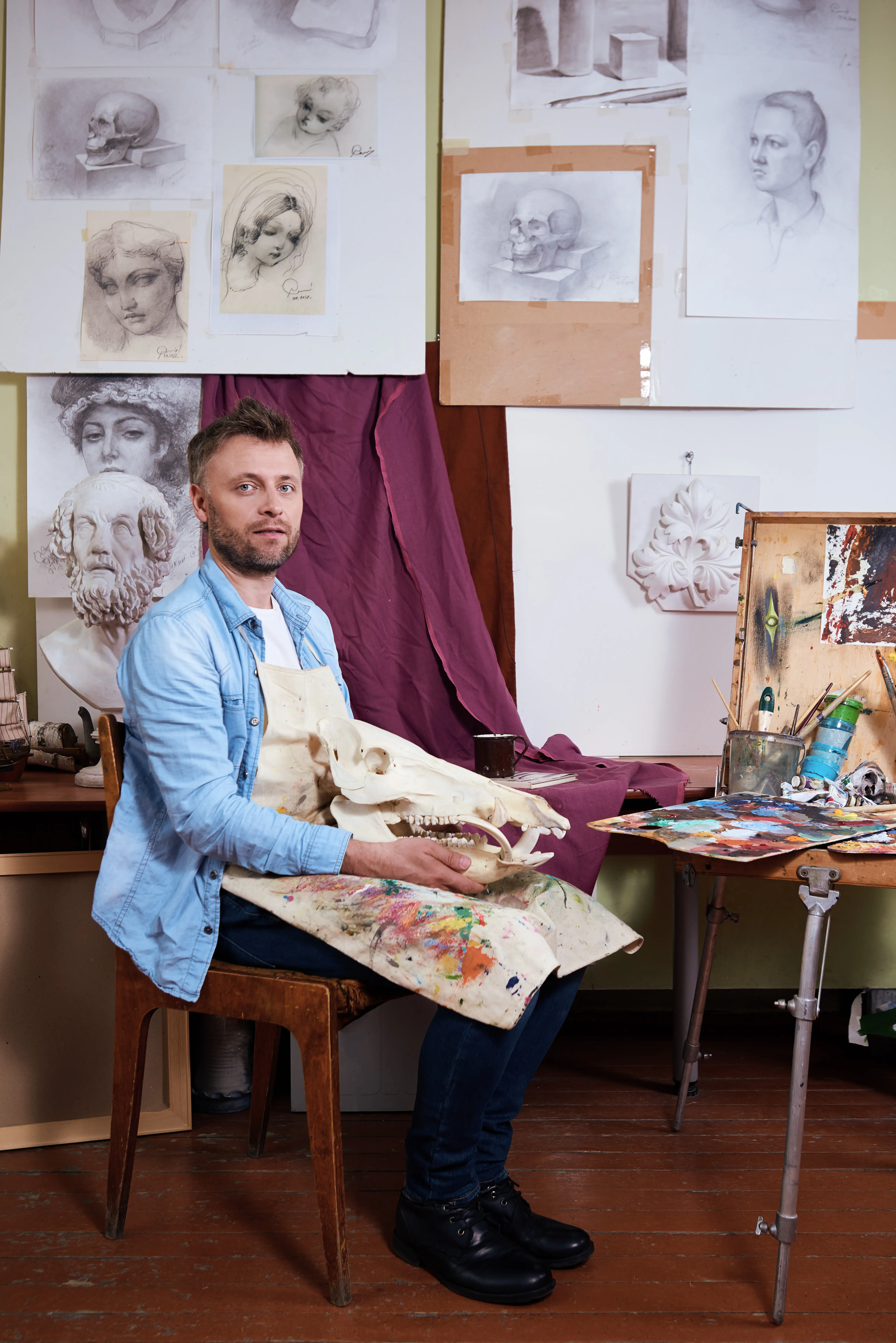 Männlicher Künstler in befleckter Schürze sitzt mit einem Dinosaurierschädel in seinem Atelier.